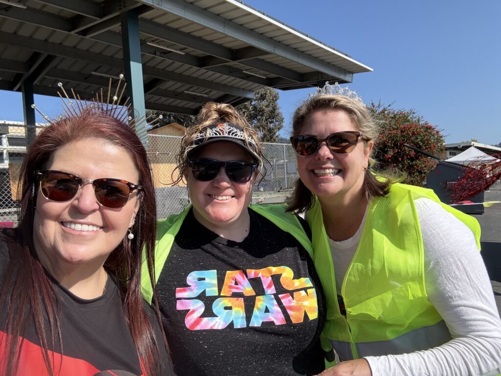Three women smiling outdoors on a sunny day, two wearing sunglasses and neon yellow safety vests, and one wearing a colorful "Star Wars" shirt and a visor. A covered structure and foliage are visible in the background.