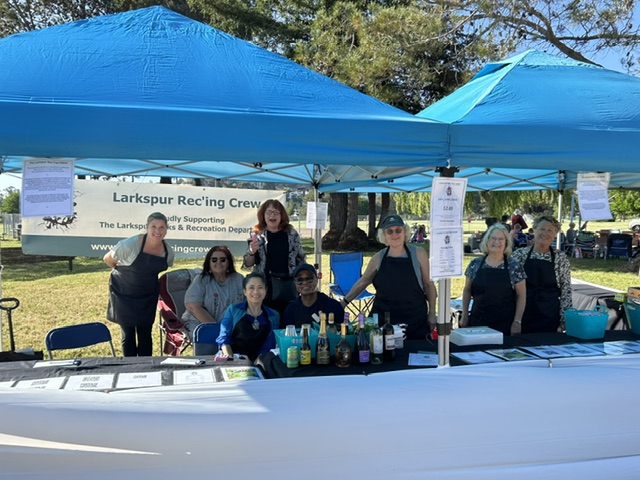 A group of people smiling and posing under blue canopy tents at an outdoor event. A banner behind them reads “Larkspur Rec’ing Crew.” They are standing behind a table with drinks and informational materials. Trees and grass are visible in the background.