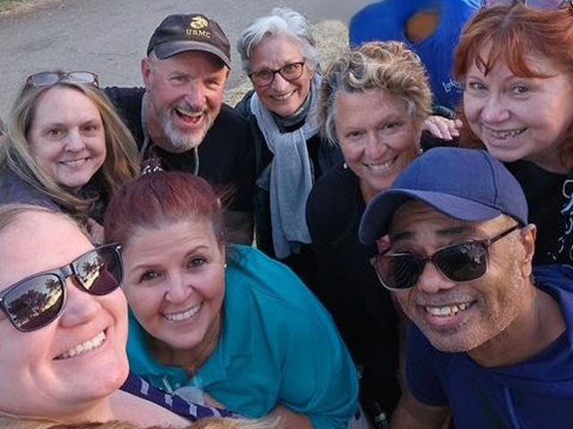 A group of nine smiling adults, both men and women, stand close together outdoors, posing for a cheerful selfie on a sunny day.